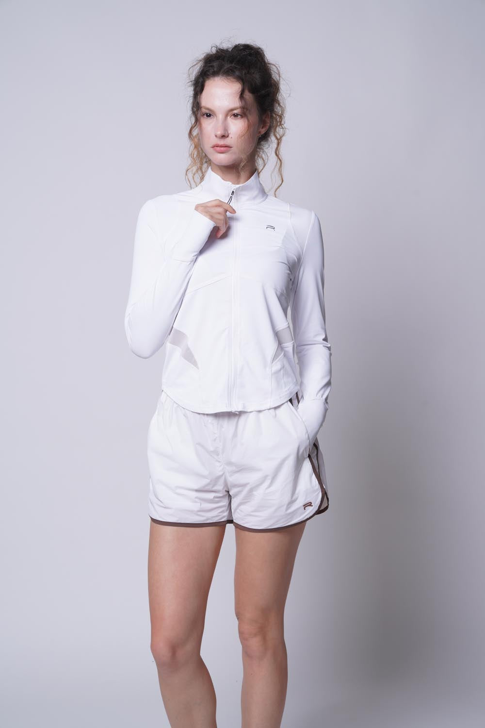 Female model wearing the white Ramsports Court Sculpt Full-Zip Jacket looking away with left hand on the pocket and her right hand holding the jacket zipper