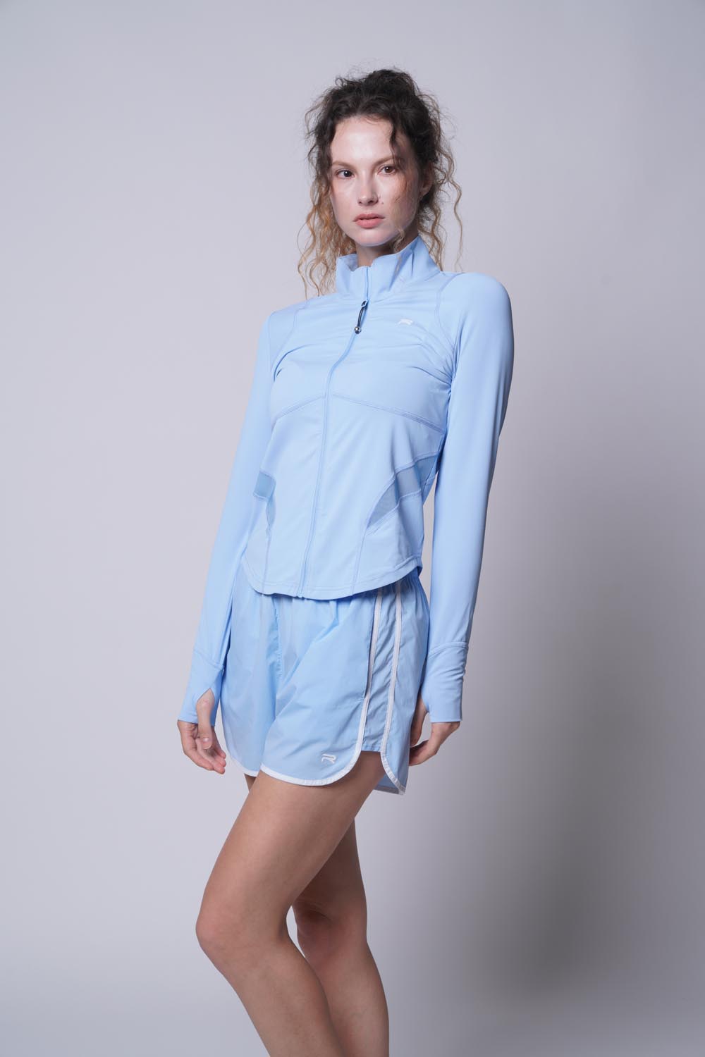 Female model wearing a blue Ramsports Court Sculpt Full-Zip Jacket with left leg bent forward, arms at her sides, looking away from the camera