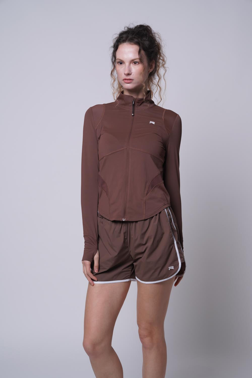 Female model wearing a brown Ramsports Court Sculpt Full-Zip Jacket standing with hands at her sides, looking away from the camera