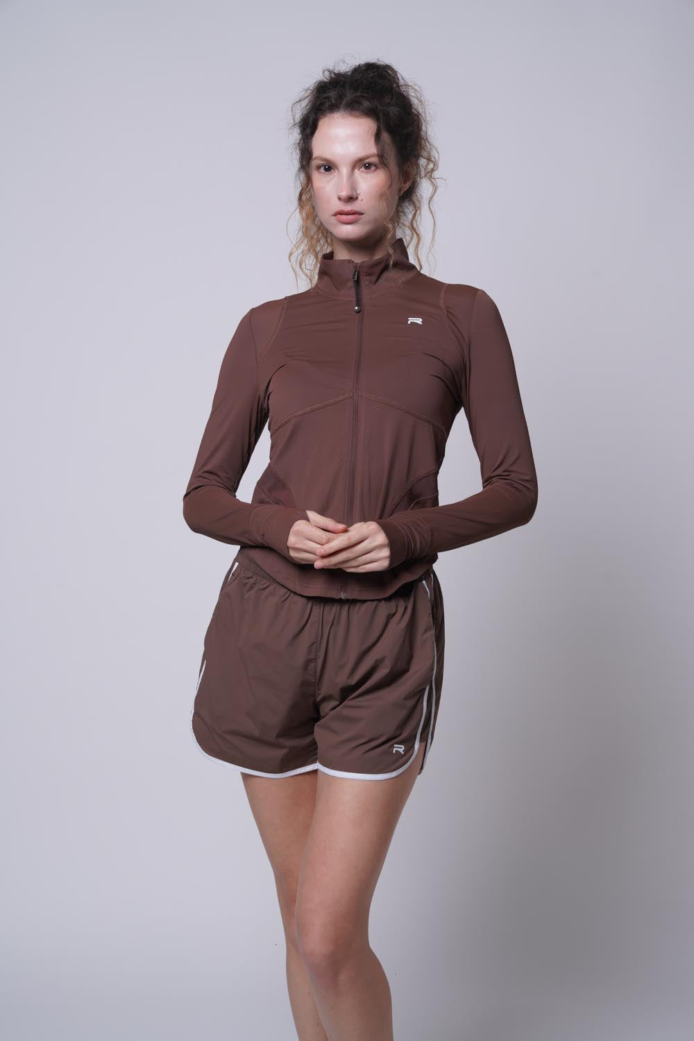 Female model wearing a brown Ramsports Court Sculpt Full-Zip Jacket and brown shorts posing modestly with her hands held together in front