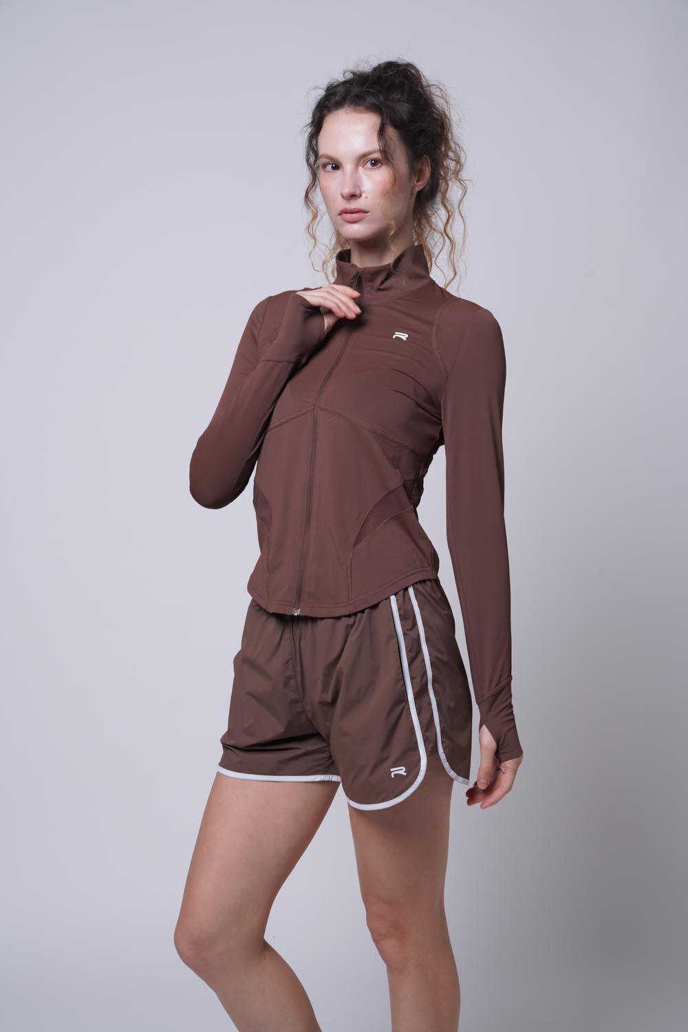 Female model wearing a brown Ramsports Court Sculpt Full-Zip Jacket paired with brown shorts holding the jacket zipper with her right hand