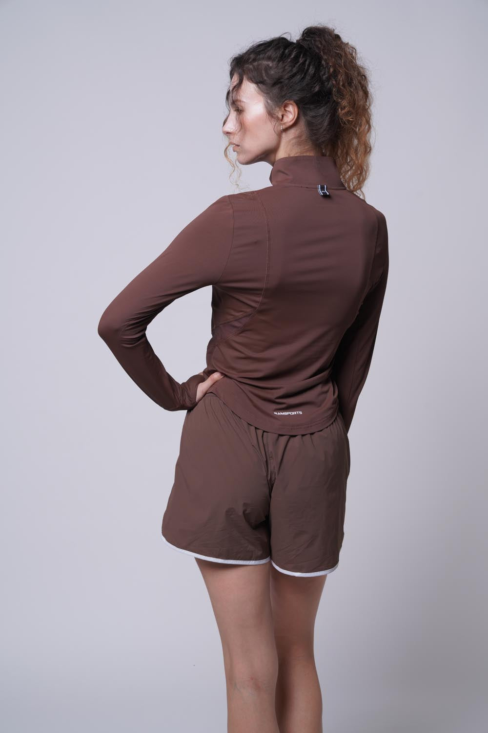 Female model wearing a brown Ramsports Court Sculpt Full-Zip Jacket with her back to the camera, left hand on her waist, head turned to the side