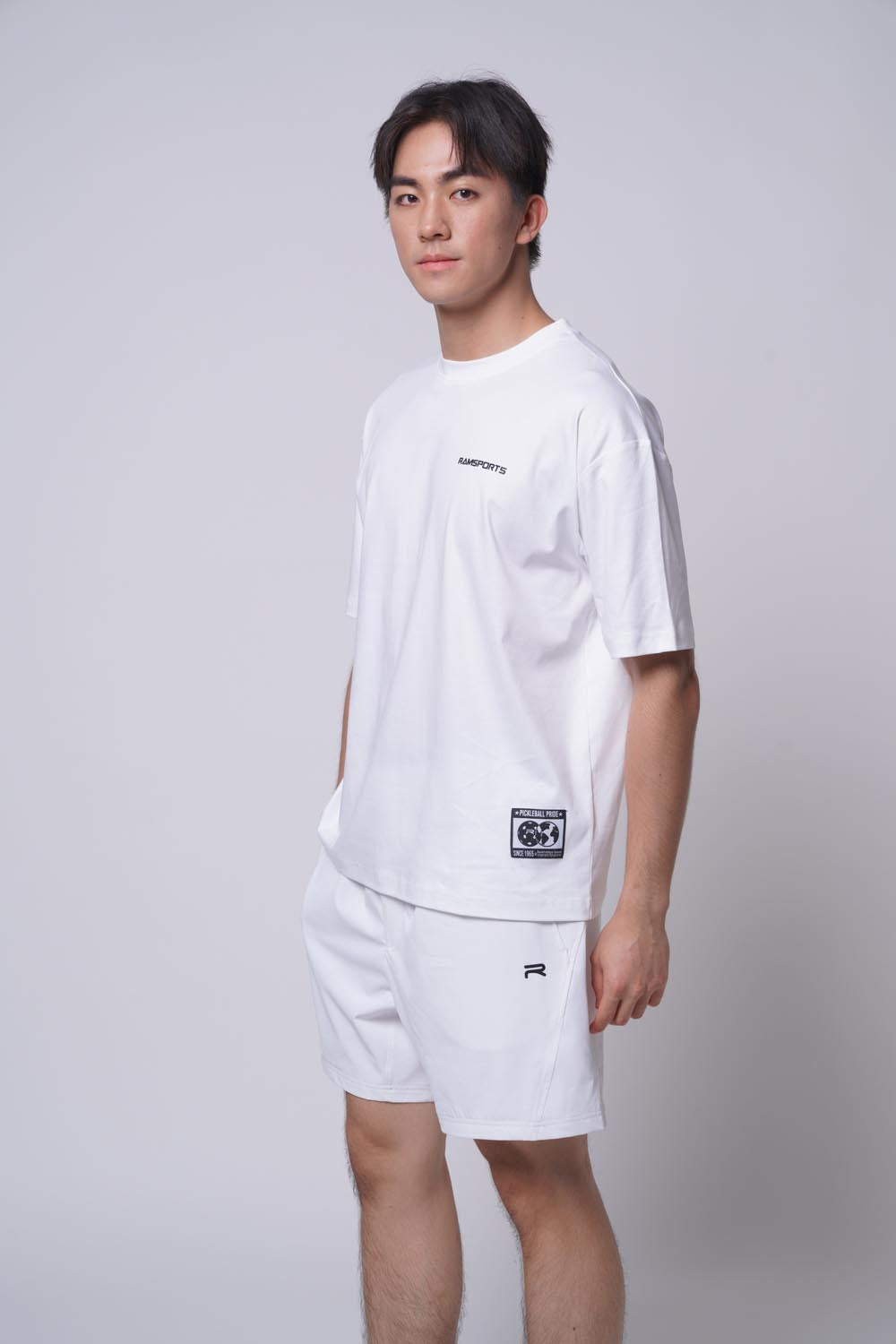 Male model wearing the white Ramsports Blueprint Back Graphic tee with white shorts against a gray background, body turned slightly to the side and looking away