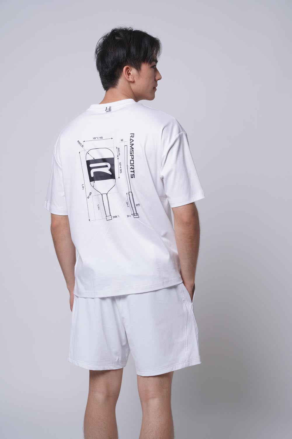 Male model displaying the back of the white Ramsports Blueprint Back Graphic tee with white shorts on a gray background