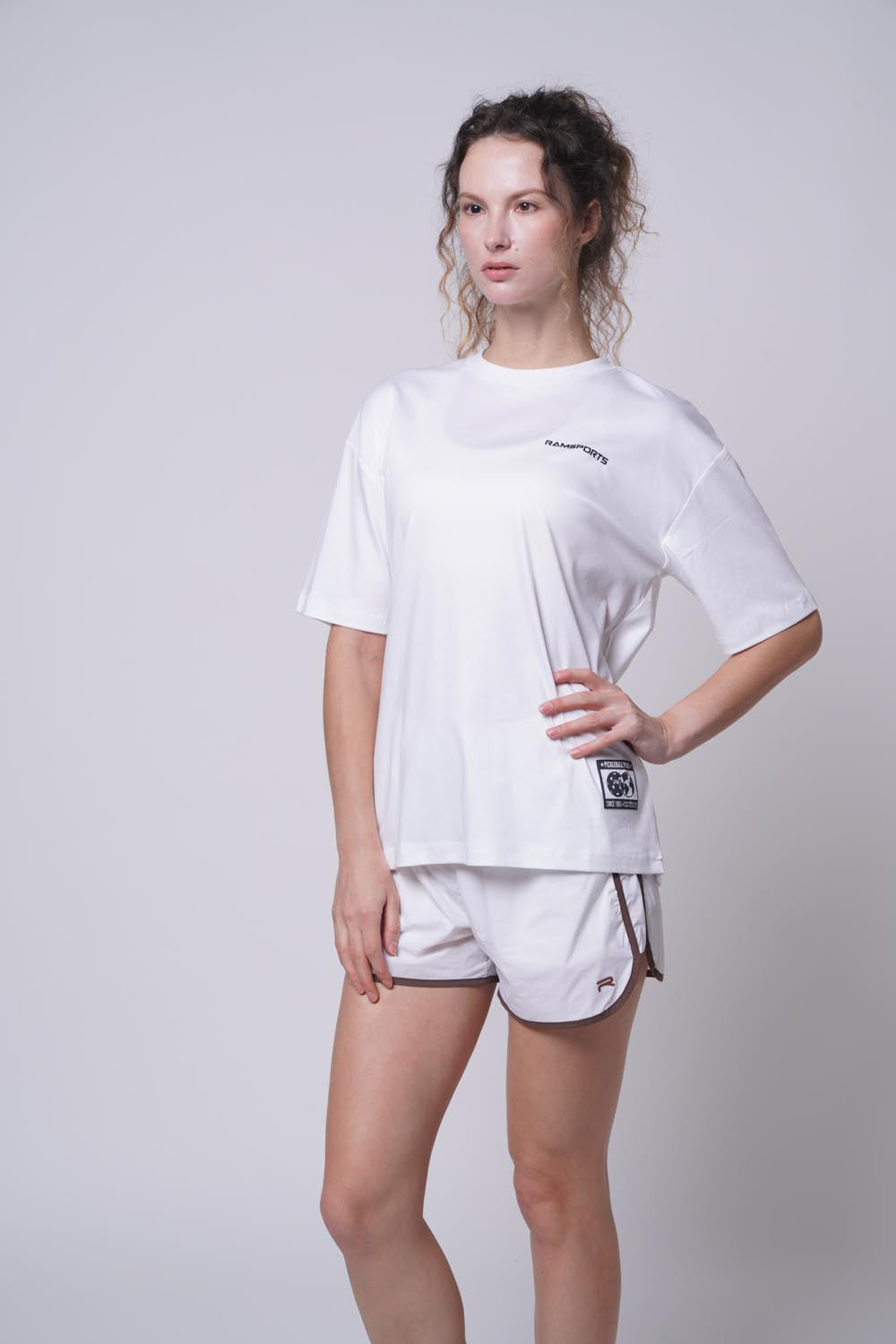 Female model in a white Ramsports Blueprint Back Graphic tee with white shorts, left hand on waist, looking away