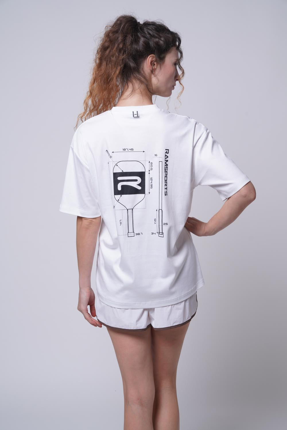 Female model displaying the back of the white Ramsports Blueprint Back Graphic tee paired with white shorts and her right hand on her waist