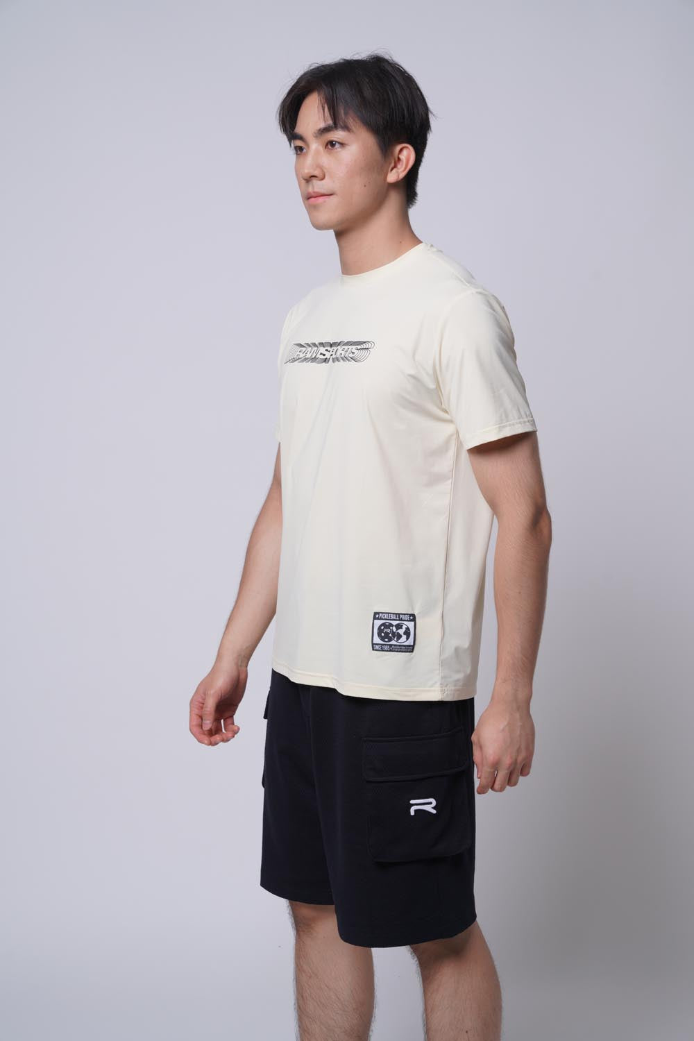 Male model wearing the beige Ramsports Playbook Court Diagram tee against a gray background, body turned slightly to the side with hands at his sides