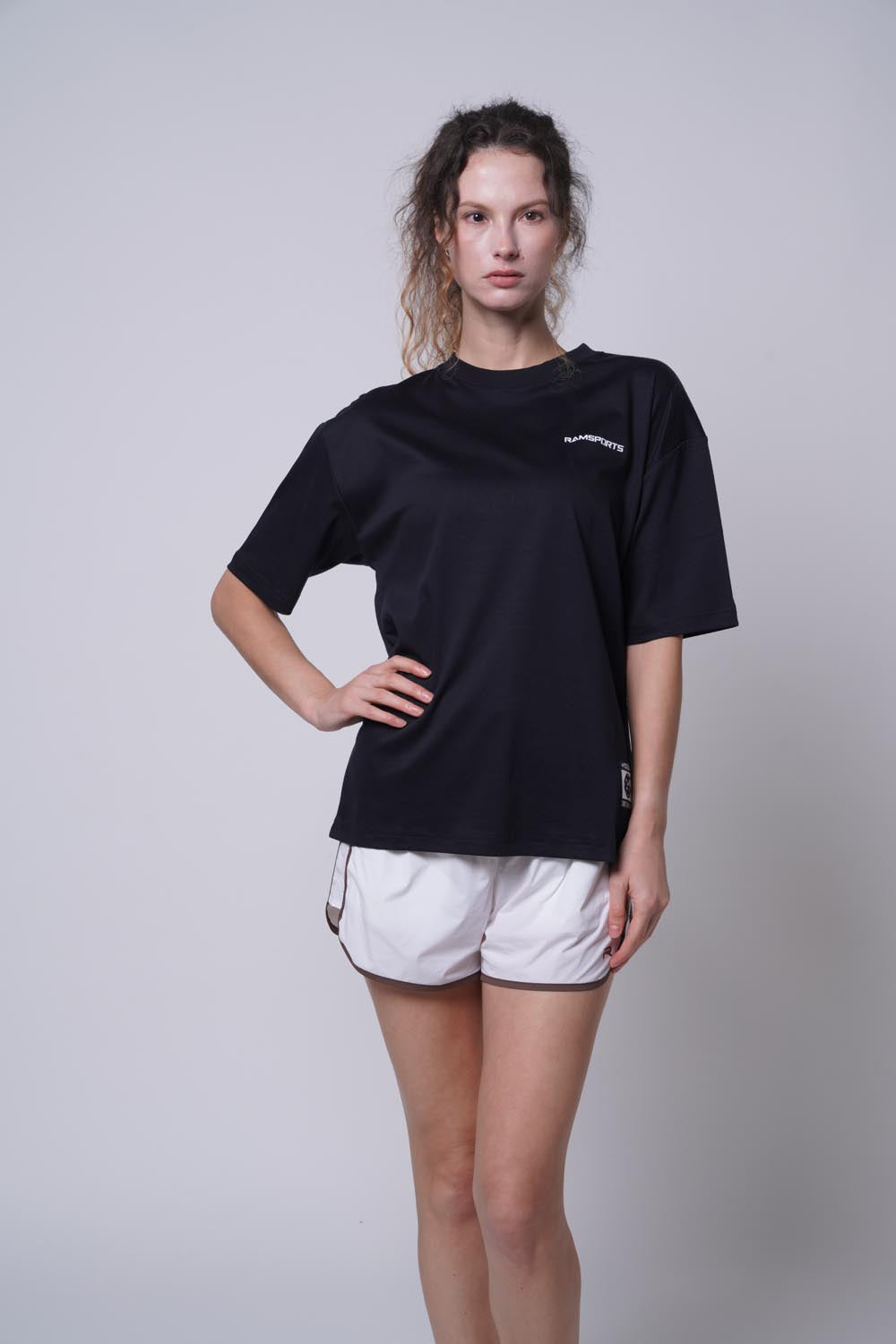 Female model wearing the black Ramsports Blueprint Back Graphic tee with white shorts against a gray background, facing the camera with one hand on her waist