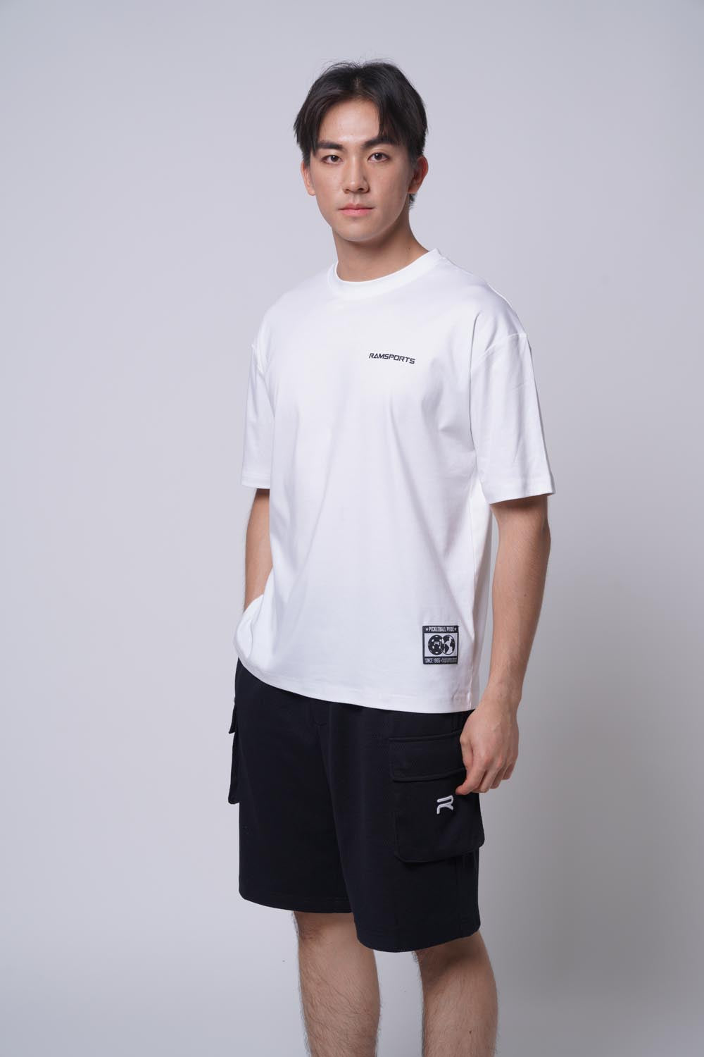 Male model posing in a white Ramsports Blueprint Back Graphic t-shirt with black shorts, facing the camera with one hand in his pocket on a gray background
