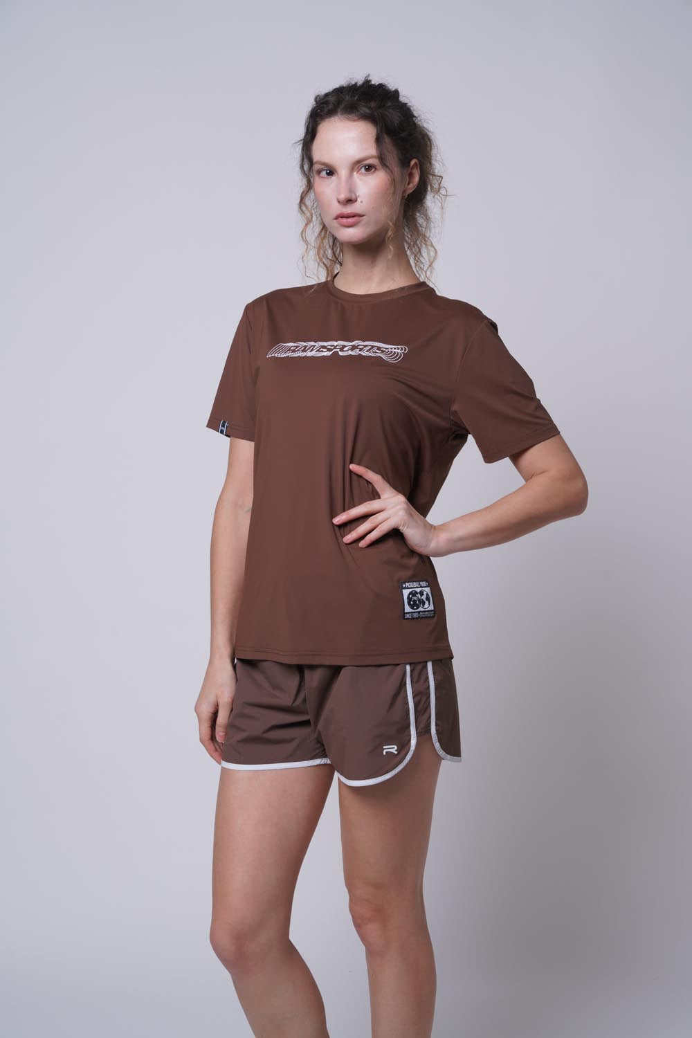 Female model posing with one hand on her waist, wearing a brown Ramsports Flow Panel Split-Hem Short.