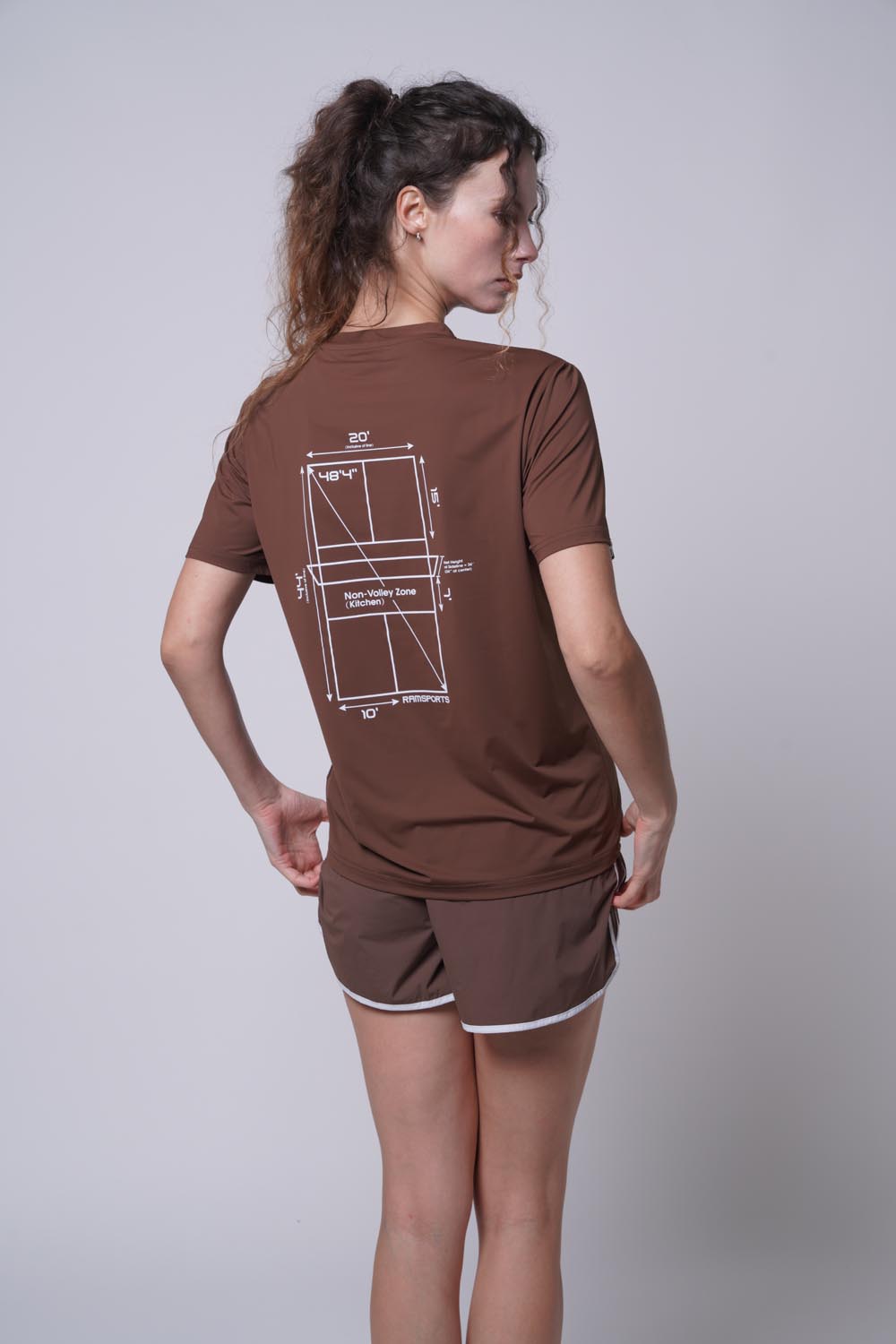 Female model displaying the back of the brown Ramsports Playbook Court Diagram Tee on a gray background