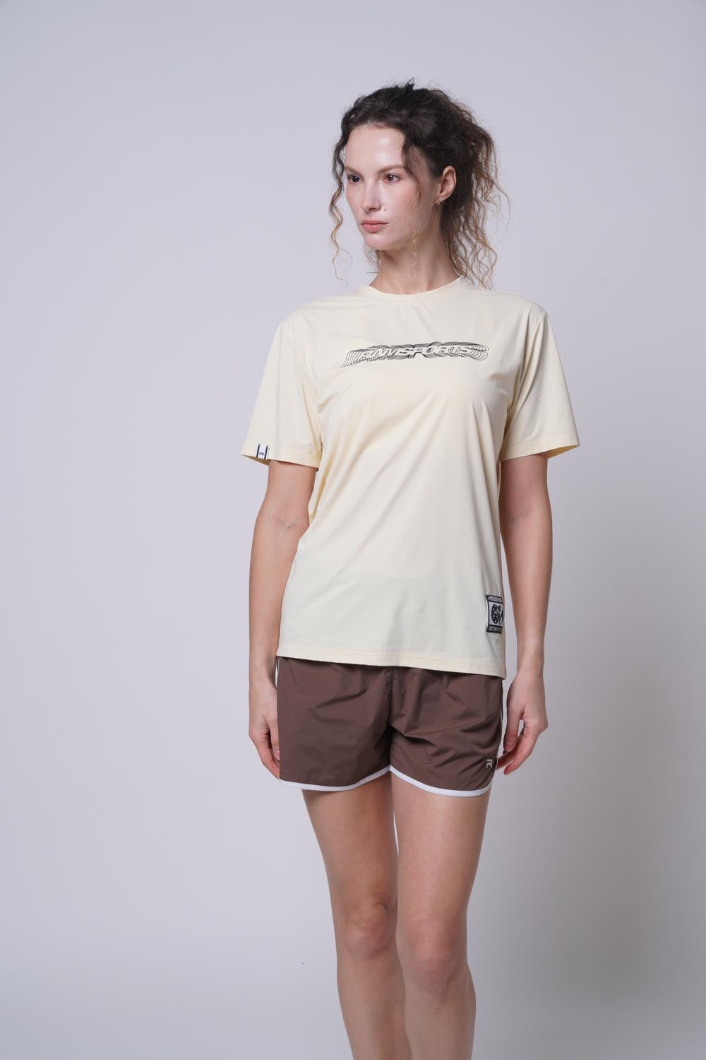 Female model wearing the beige Ramsports Playbook Court Diagram tee with brown shorts against a gray background, looking away from the camera