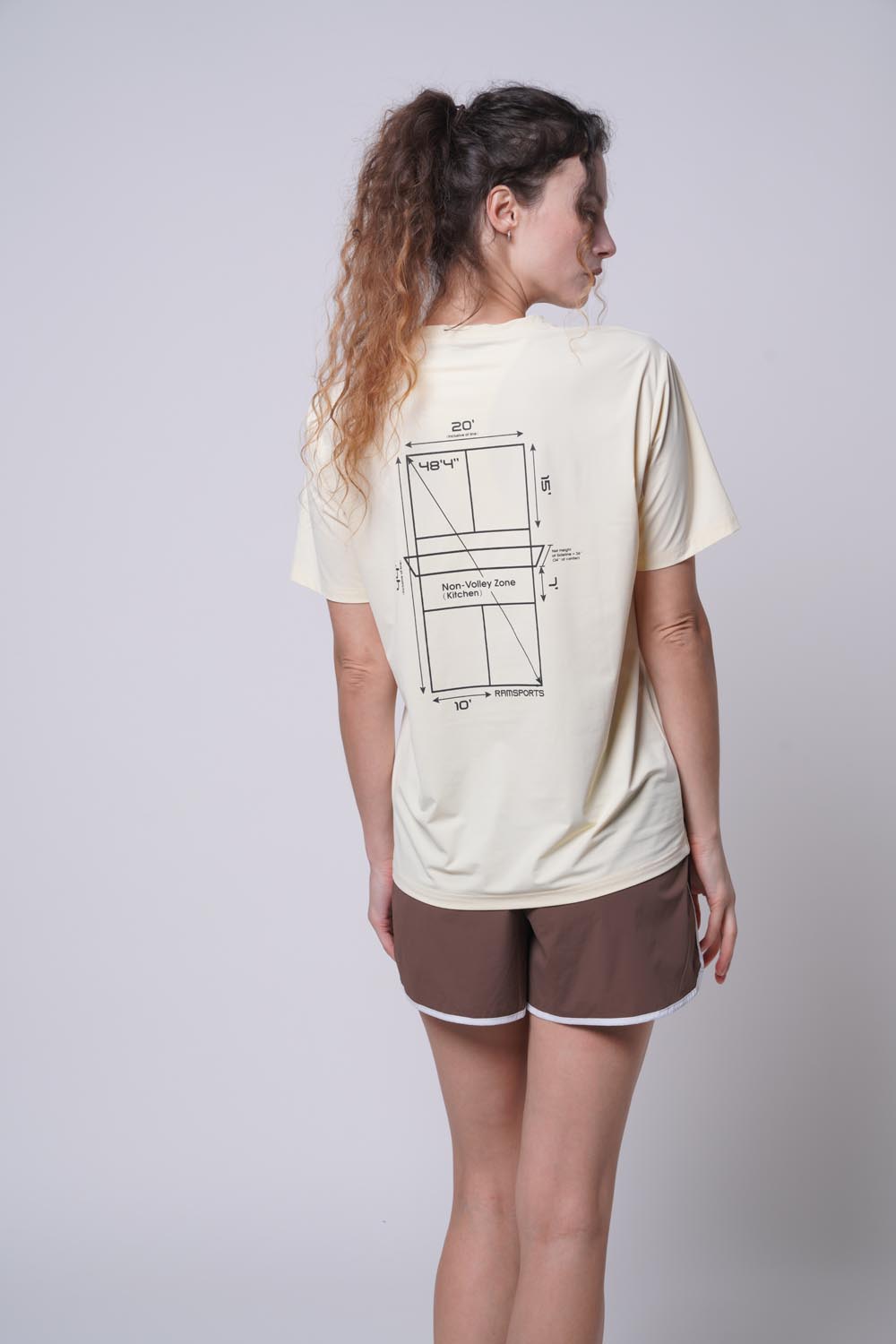 Back view of female model wearing a beige Ramsports Playbook Court Diagram tee with brown shorts
