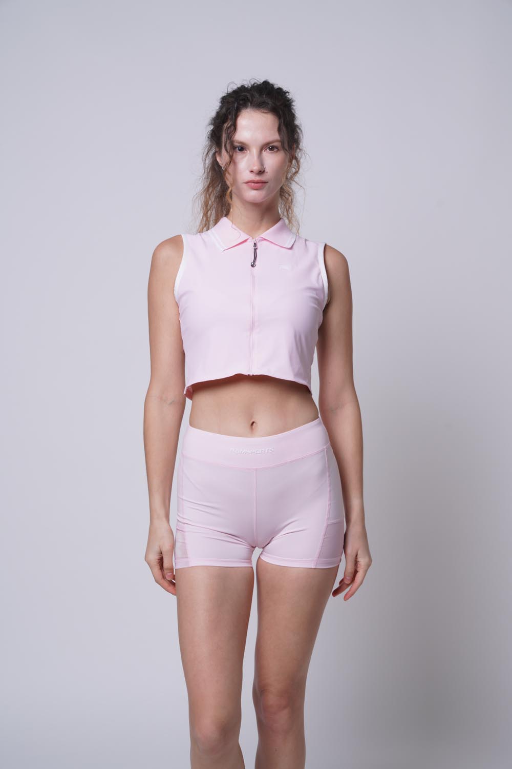 Female model modeling a pink Ramsports Serve Zip Fitted Shorts set, facing the camera with hands at her sides and gaze forward.