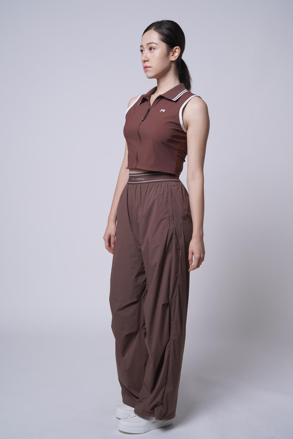 Full body picture of Asian female model posing sideways while wearing Ramsports serve zip crop polo in brown