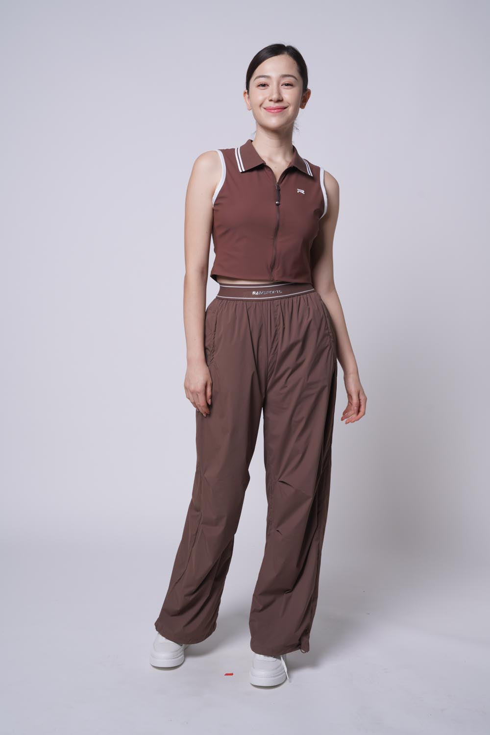 Asian female model in Ramsports Serve Zip Crop Polo and brown Court Sculpt Track Pants, smiling at the camera.