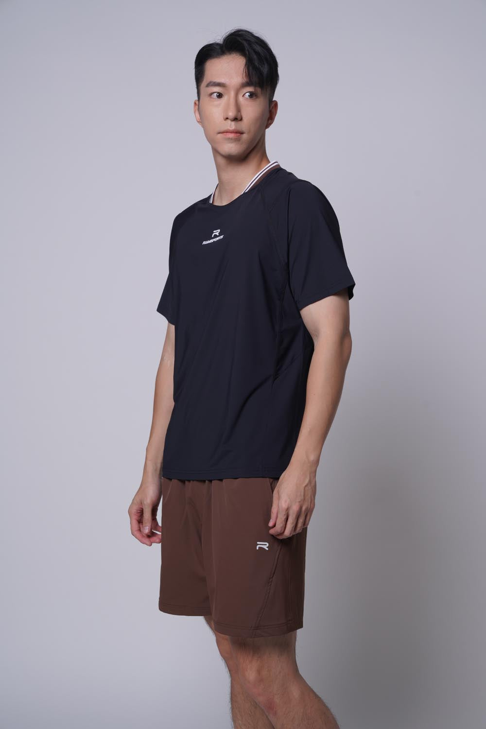 Asian male model facing sideways, wearing ramsports Edge Line Raglan Tee in black with brown shorts