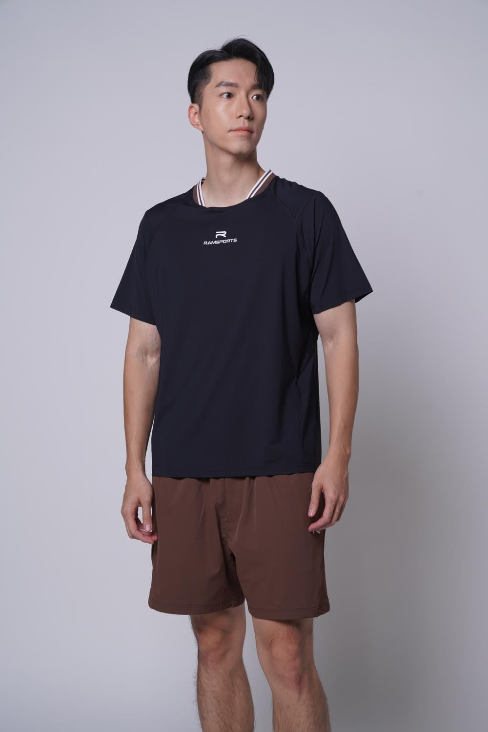 Asian male model looking away at the camera, wearing ramsports Edge Line Raglan Tee in black with brown shorts