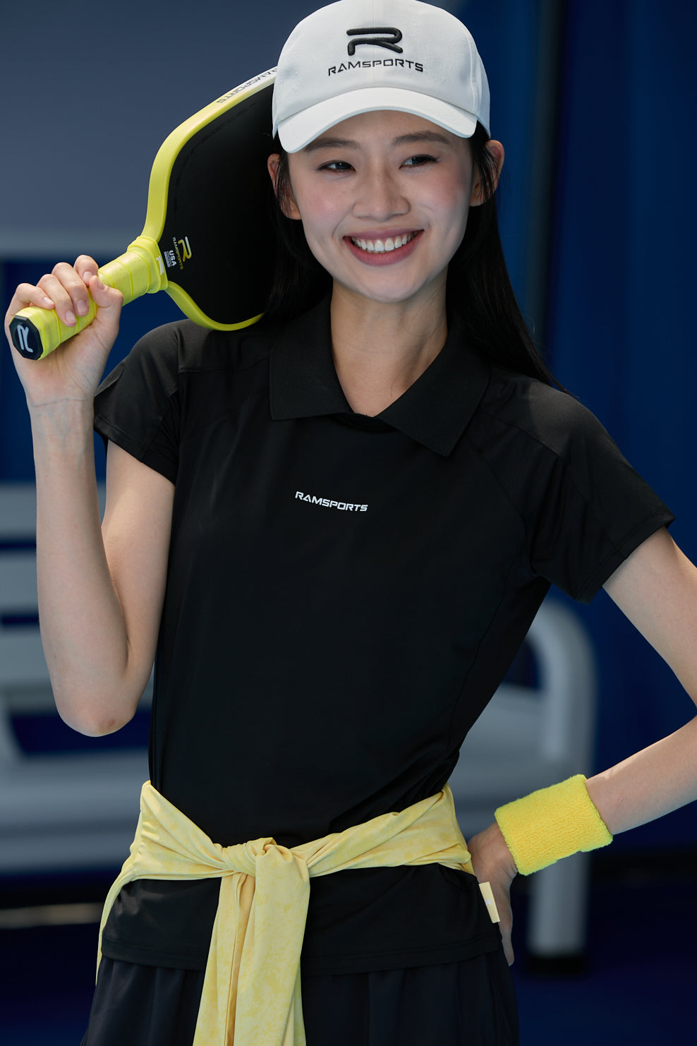 Asian female model wearing an all-black outfit with Ramsports AirFlow Mesh Polo and black shorts, holding a paddle over her shoulder