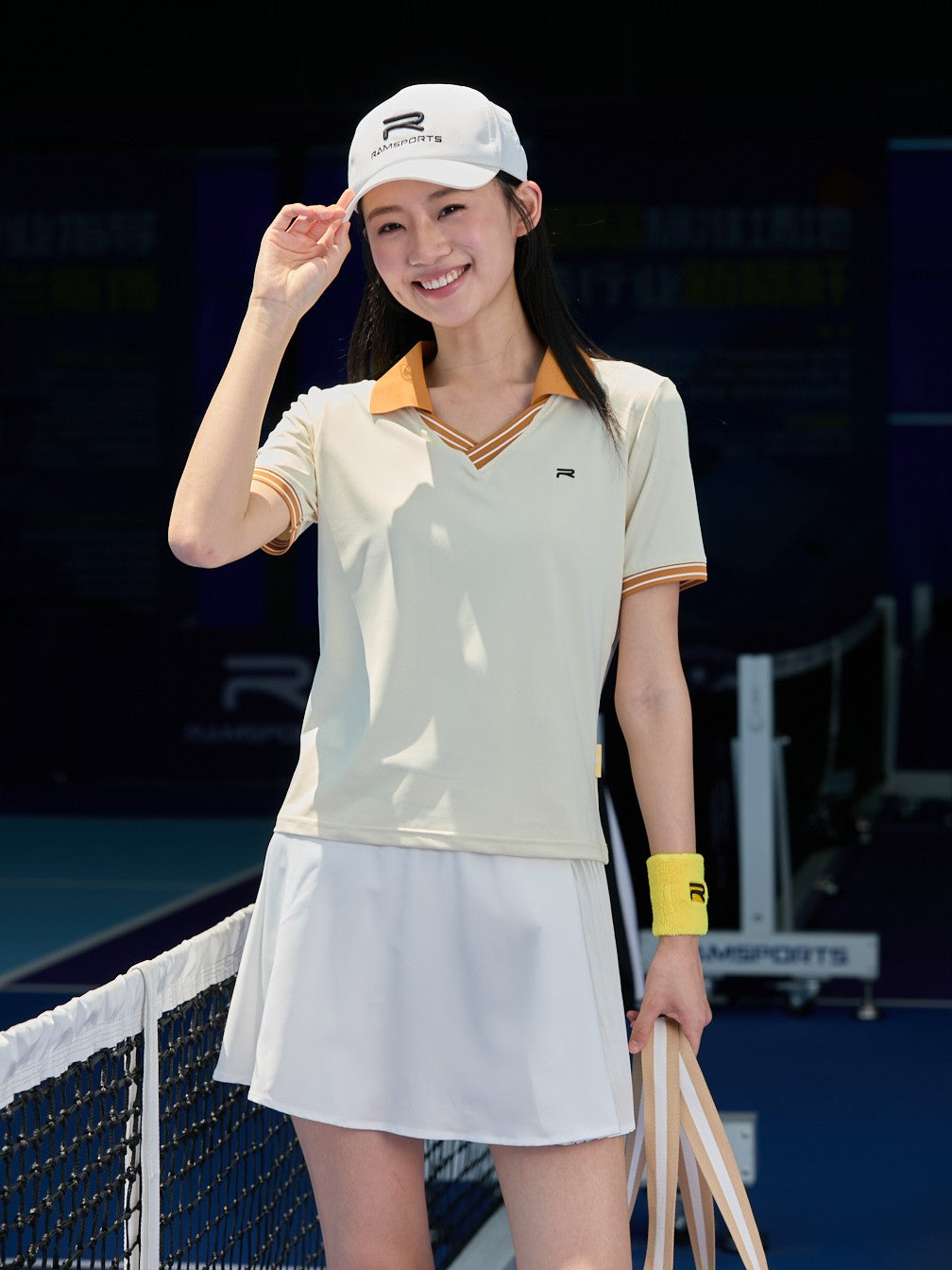 Smiling Asian female model wearing a white Ramsports AirFlow Pleated Skort and a light yellow shirt with orange collars