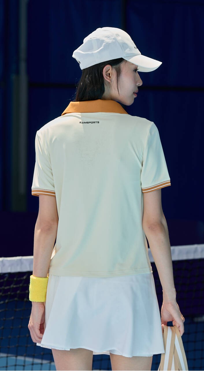 Smiling Asian female model turning away from the camera, wearing a white Ramsports AirFlow Pleated Skort and a light yellow shirt with orange collar