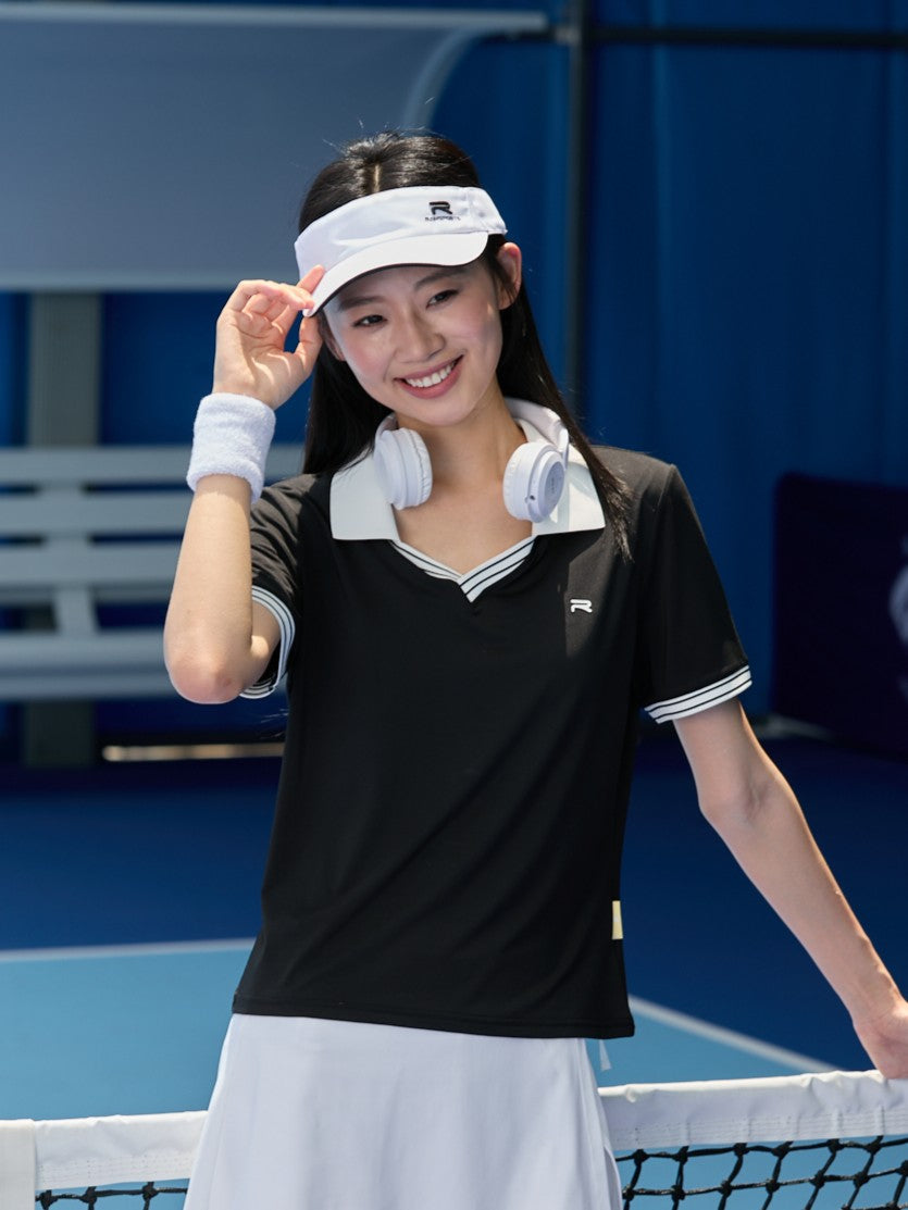 Smiling Asian female model wearing a black Ramsports AirFlow V-Neck Polo and a white skort, holding the brim of the cap she's wearing