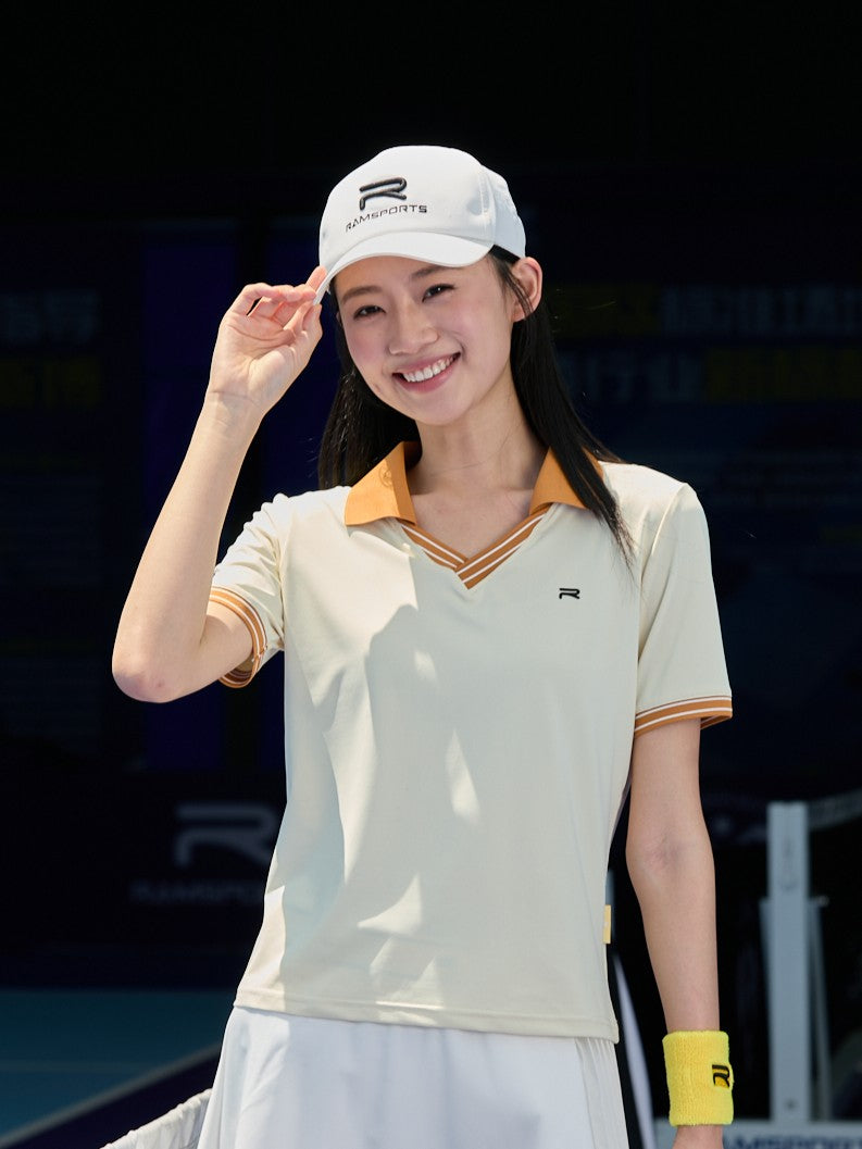 Asian female model wearing a khaki Ramsports AirFlow V-Neck Polo and a white skort, holding the brim of her cap
