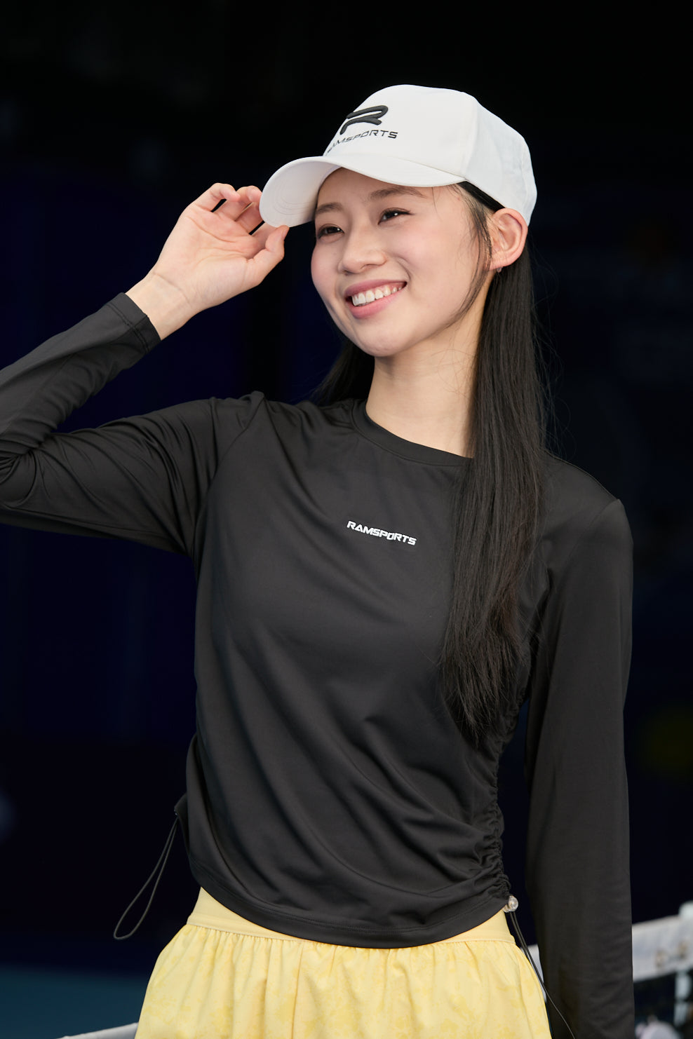 Asian female model wearing a black Ramsports CoolDry Long-Sleeves Tee and yellow shorts, with one hand holding the visor of her cap