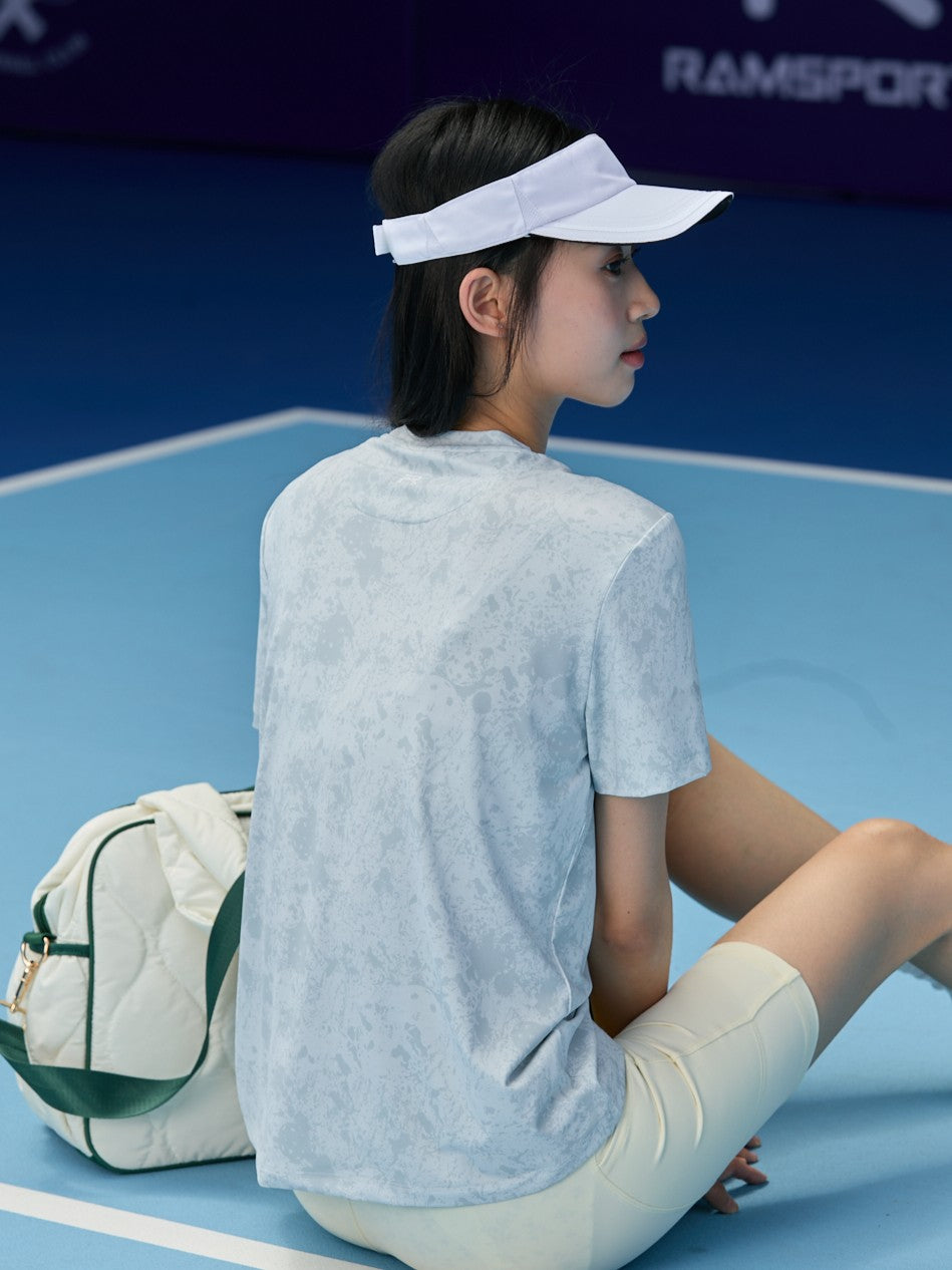 Asian female model facing away, wearing a gray Ramsports CoolDry Marble Print Tee, light yellow shorts, a white visor, and a yellow wristband