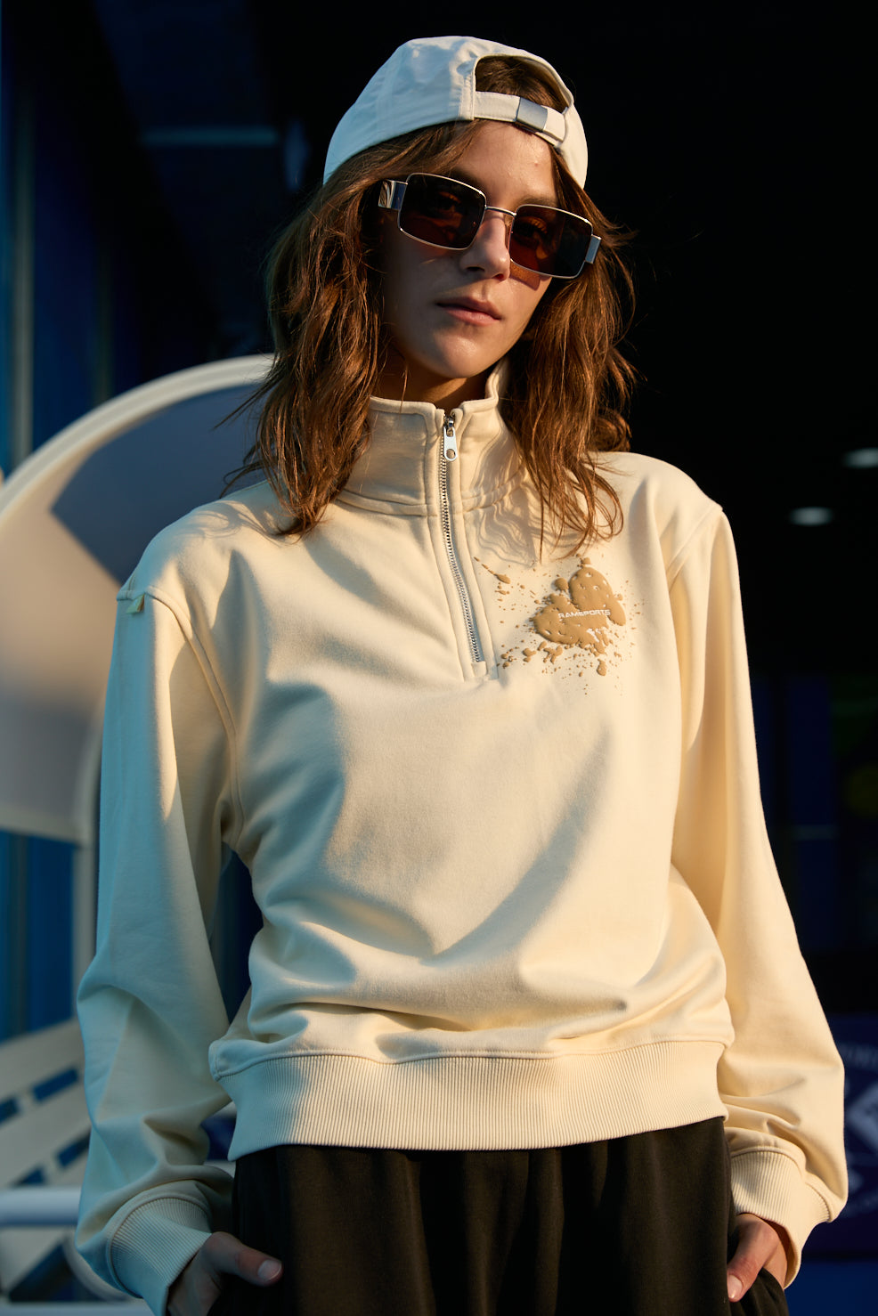 Female model wearing a khaki Ramsports EcoSoft Half Zip Logo Sweatshirt with black bottoms, a white cap, and sunglasses, with her hands in her bottoms' pockets