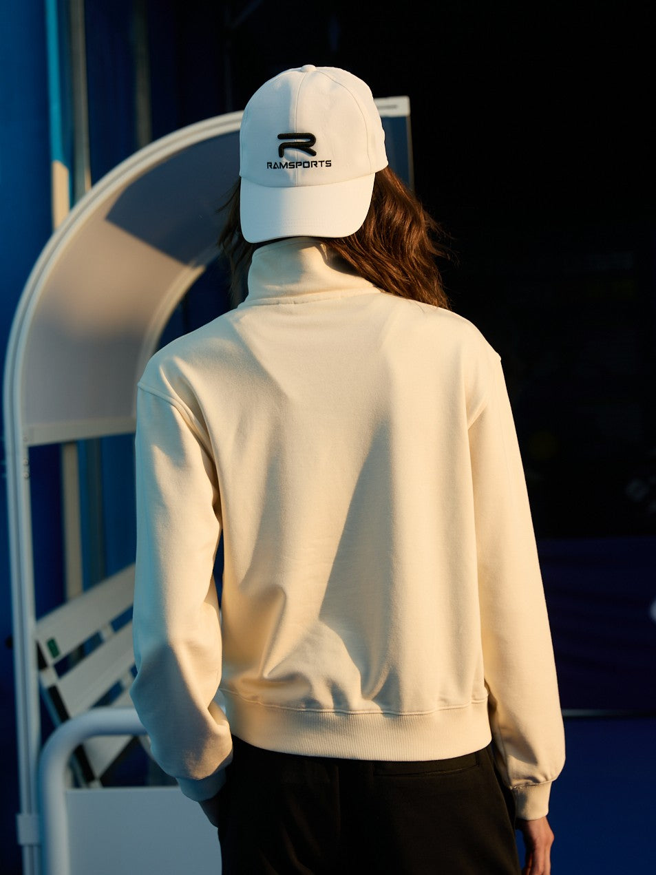 Female model turned away, wearing a khaki Ramsports EcoSoft Half Zip Logo Sweatshirt with black bottoms and a white cap.