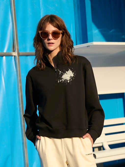 Female model wearing a black Ramsports EcoSoft Half Zip Logo Sweatshirt and khaki bottoms with her hands on her pockets