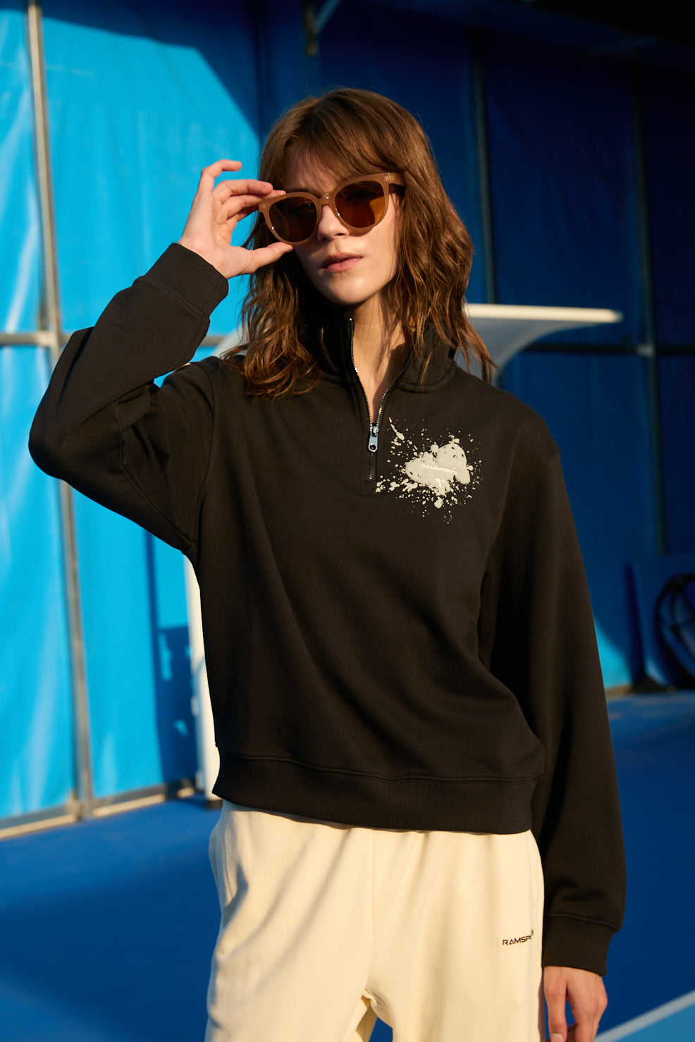 Female model wearing a black Ramsports EcoSoft Half Zip Logo Sweatshirt and khaki bottoms as she's holding her sunglasses