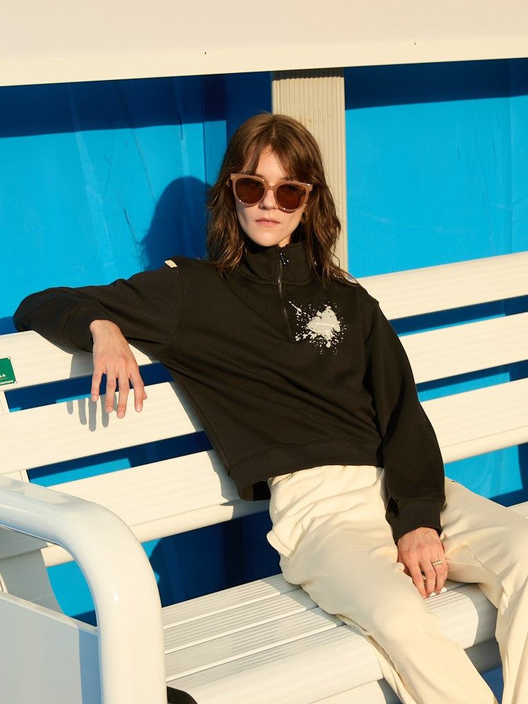 Female model wearing a black Ramsports EcoSoft Half Zip Logo Sweatshirt and khaki bottoms as she's sitting down on a white bench