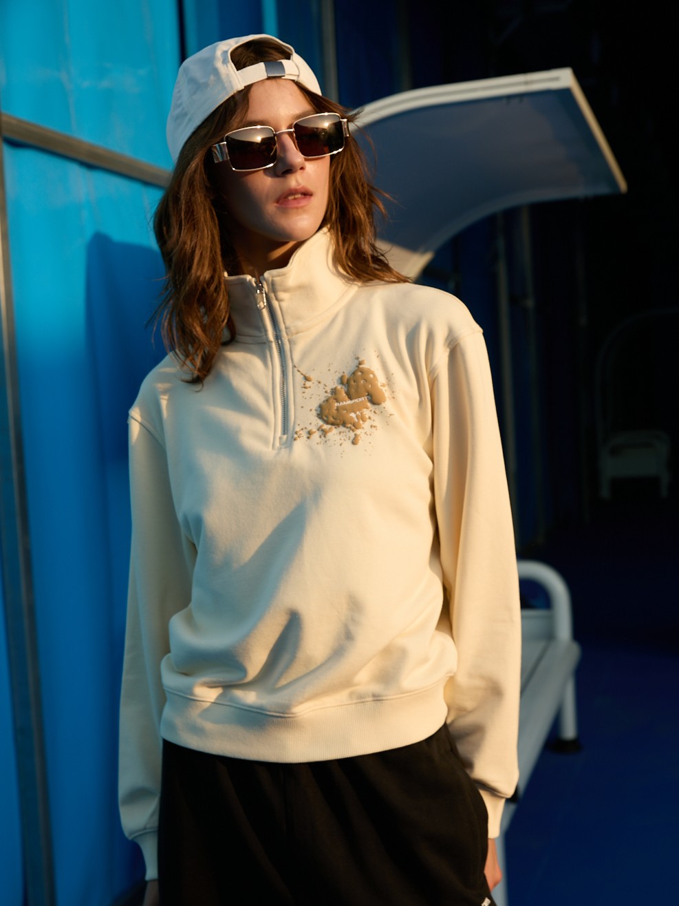 Female model wearing a khaki Ramsports EcoSoft Half Zip Logo Sweatshirt paired with black bottoms, a white cap, and sunglasses as she's looking away