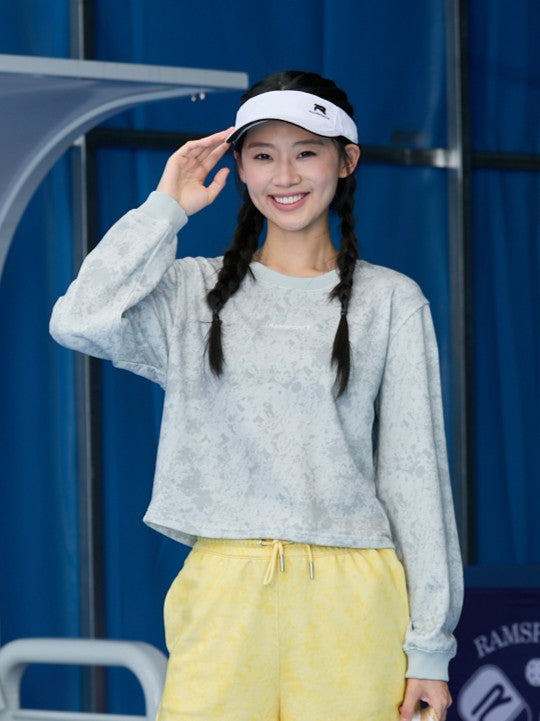 Smiling Asian female model standing, wearing a gray Ramsports EcoSoft Marble Print Crop Sweatshirt and yellow shorts, with one hand touching her white cap
