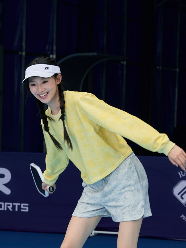 Smiling Asian female model wearing a yellow Ramsports EcoSoft Marble Print Crop Sweatshirt and gray shorts, posing as if playing pickleball while holding a paddle
