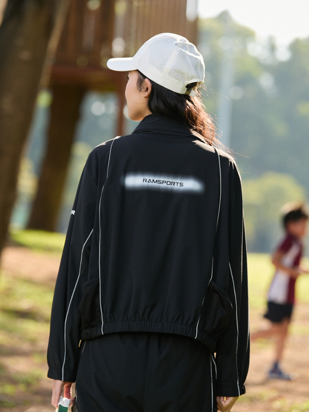 Asian female model facing backward, wearing a black Ramsports FlexTek Block Jacket with a visible white Ramsports logo on the back
