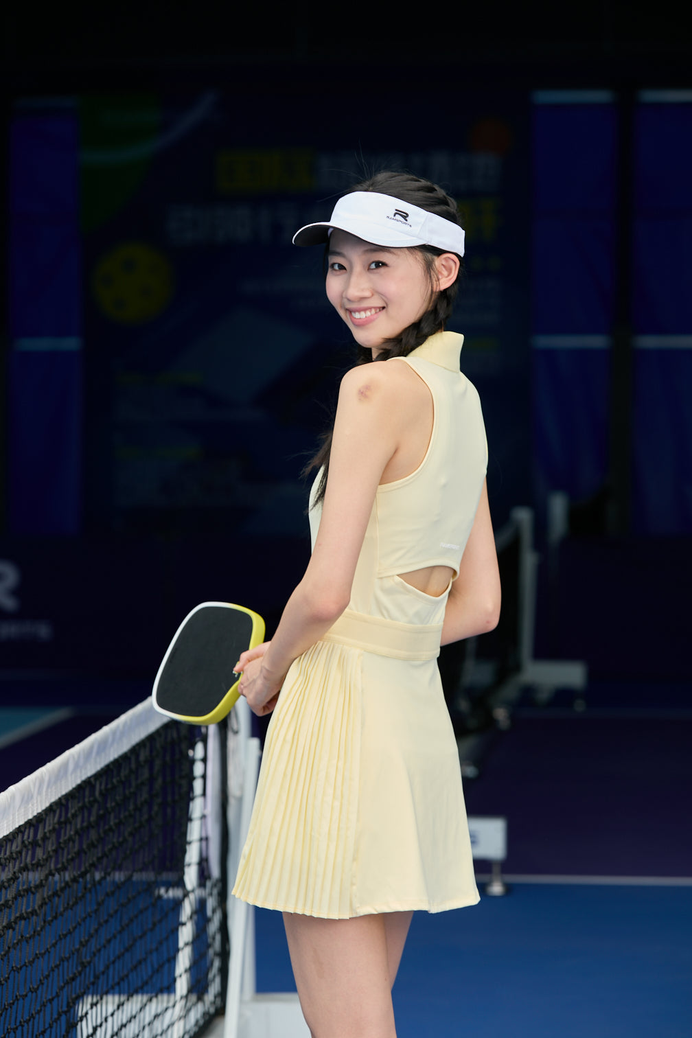 Asian female model wearing a yellow Ramsports FlexTek Court Dress with her hair in a ponytail, posing sideways near the net while holding a paddle in one hand