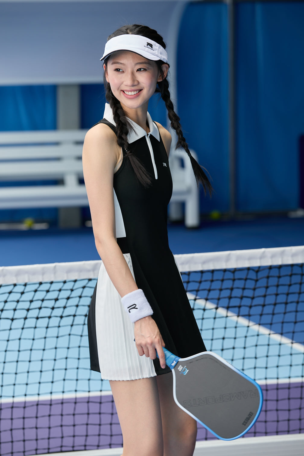 Asian female model wearing a black Ramsports FlexTek Court Dress with her hair in a ponytail, holding a paddle in one hand