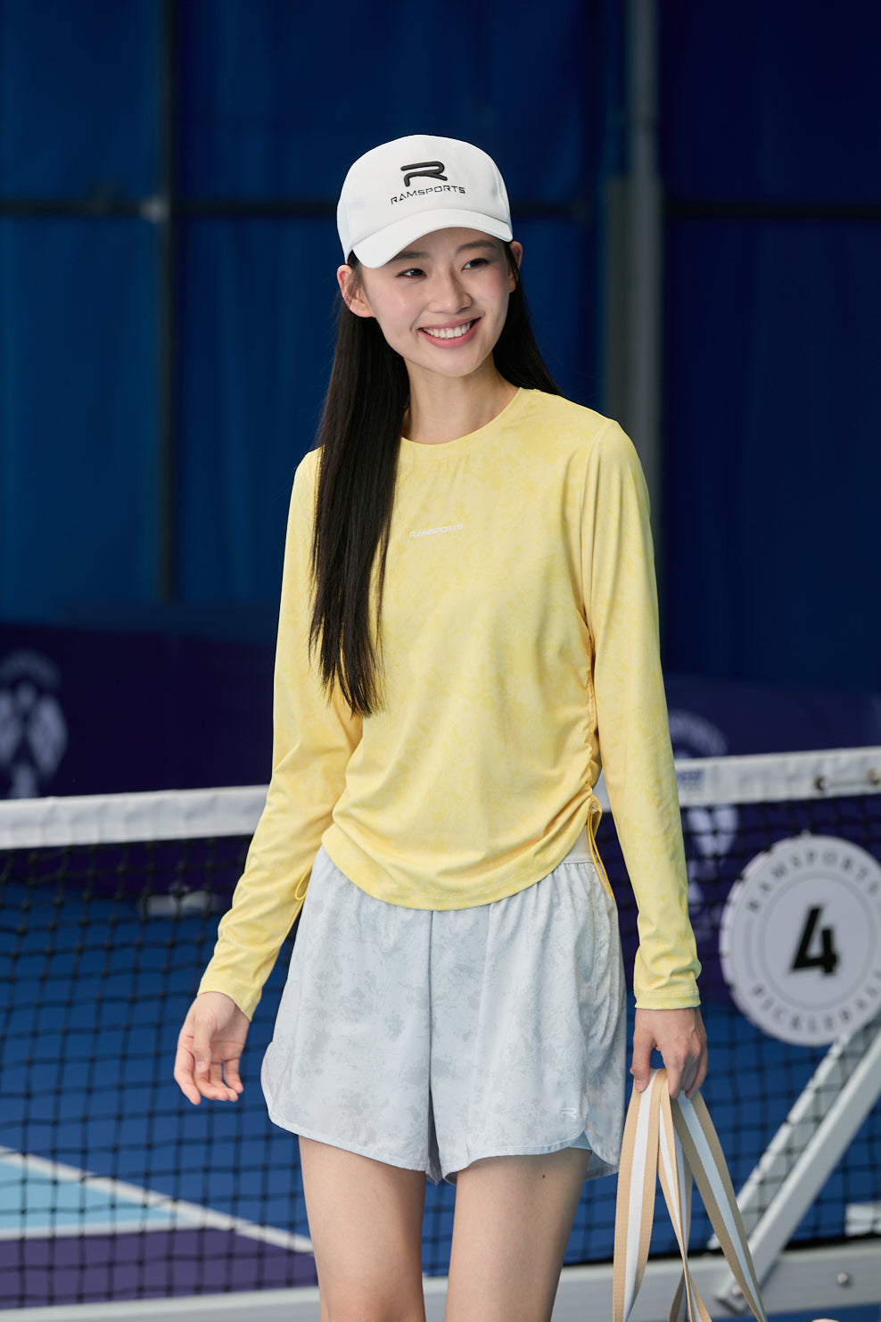 smiling asian model wearing a Ramsports cap, yellow long-sleeve top, and gray Ramsports FlexTek Marble Print Shorts