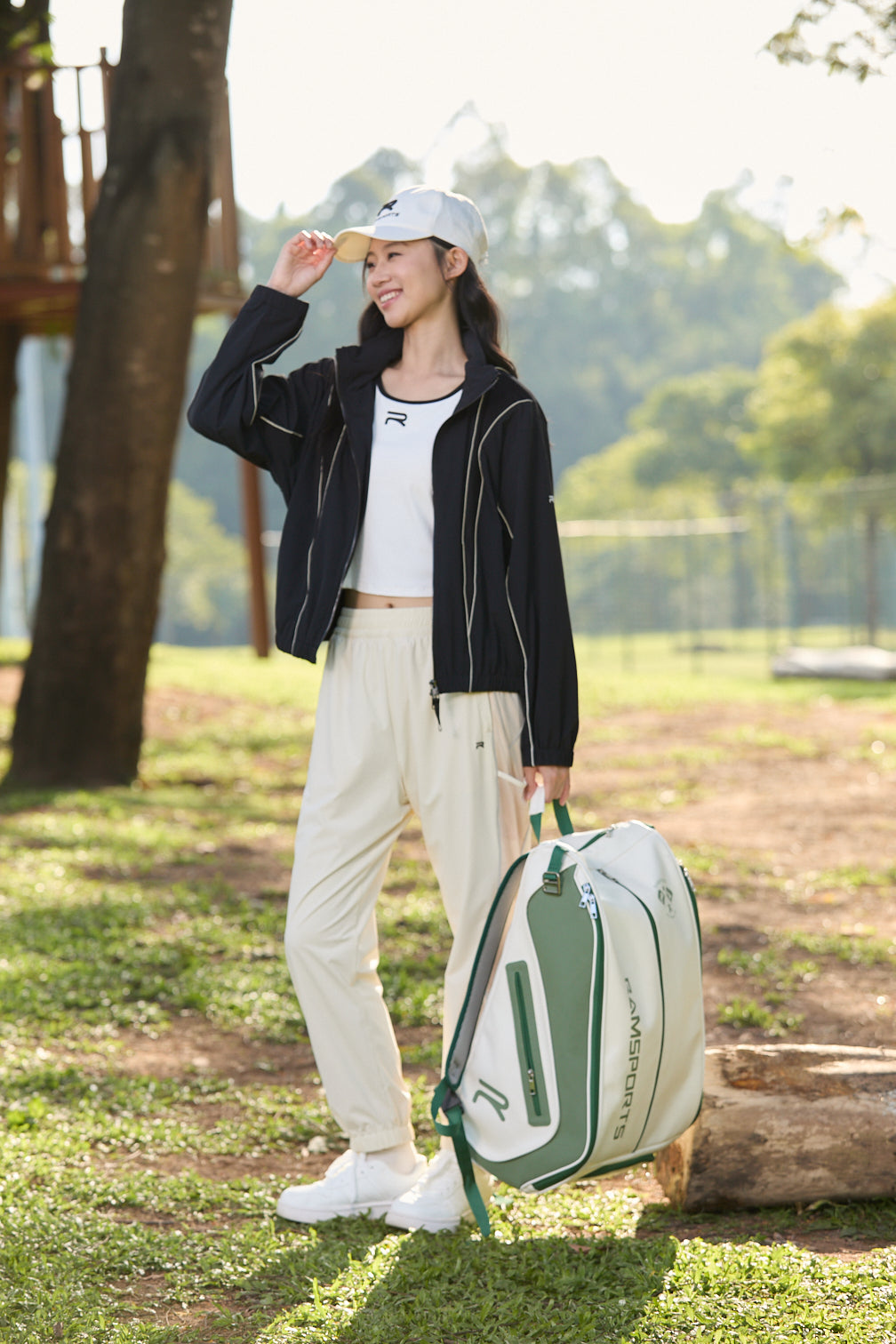 Smiling Asian female model in khaki Ramsports FlexTek Motion Joggers, holding a white cap in one hand and a backpack in the other