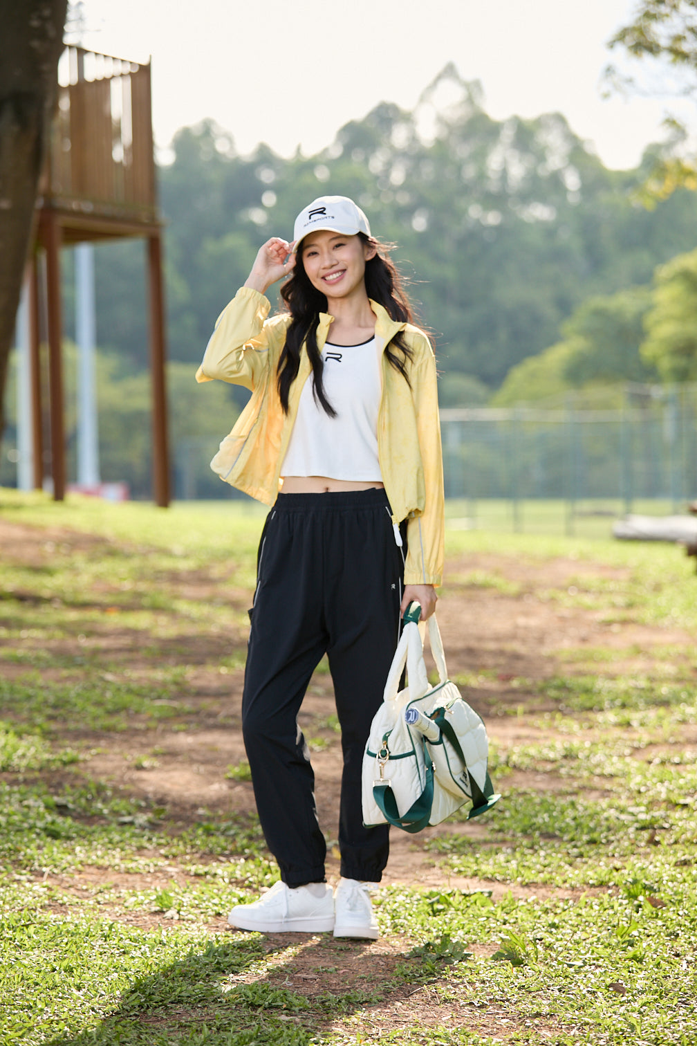 Smiling Asian female model in black Ramsports FlexTek Motion Joggers, white top, and yellow jacket, holding her white cap in her head