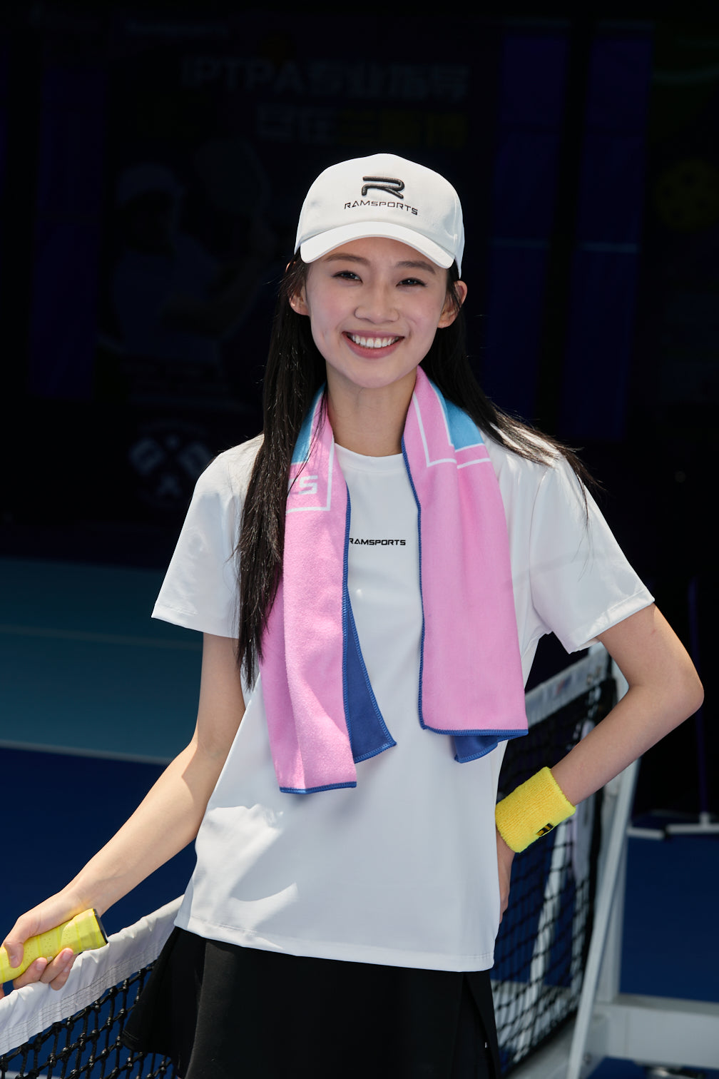 Smiling Asian female model wearing a white Ramsports FlexTek Motion Tee and a black skort, posing by the net