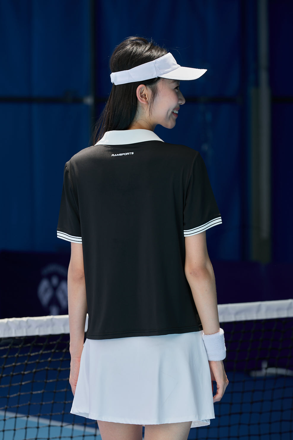 Asian female model facing away from the camera, wearing a white Ramsports FlexTek Sport Skort and black shirt, with a visor on