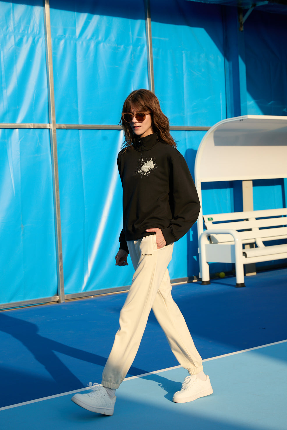 Female model in motion, wearing khaki Ramsports EcoSoft Everyday Joggers, a black jacket, and sunglasses, posed as if walking