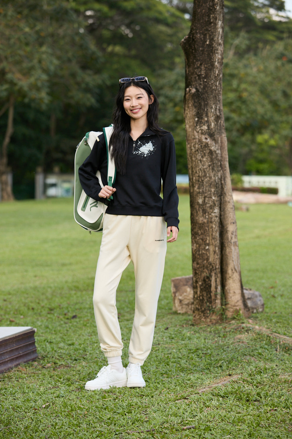 Smiling Asian female model wearing a khaki Ramsports EcoSoft Everyday Jogger and a black long-sleeve top, with one strap of a backpack over her shoulder