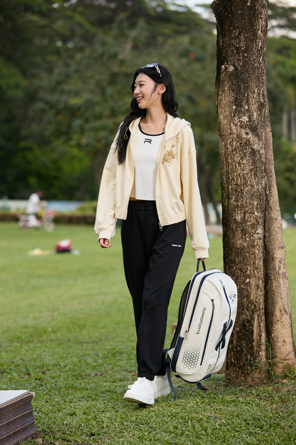 Smiling Asian female model wearing black Ramsports EcoSoft Everyday Joggers, a yellow jacket over a white top, holding a backpack in one hand