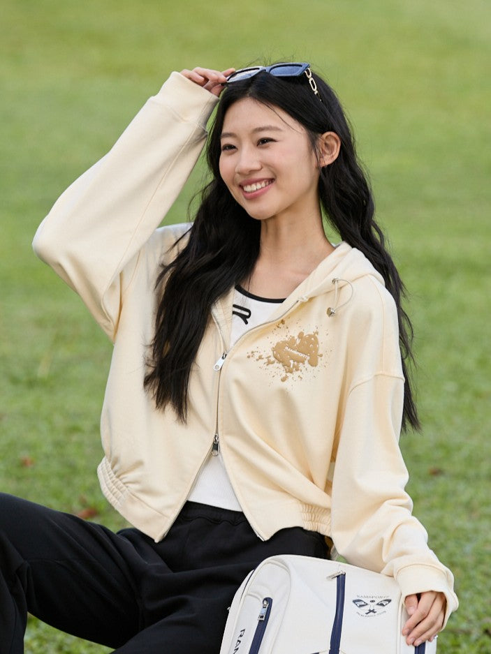 Smiling Asian female model sitting on the grass, wearing a khaki Ramsports EcoSoft Logo Hoodie over a white top