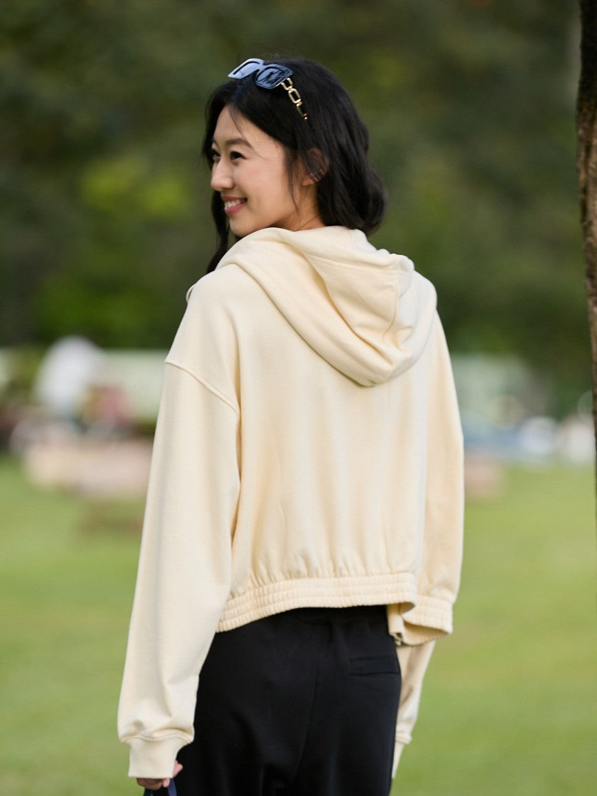 Smiling Asian female model standing slightly sideways, wearing a khaki Ramsports EcoSoft Logo Hoodie with black pants